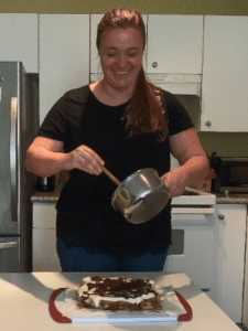 A young woman in a black shirt smiling as she holds a pot and spoon over a tray of desserts.