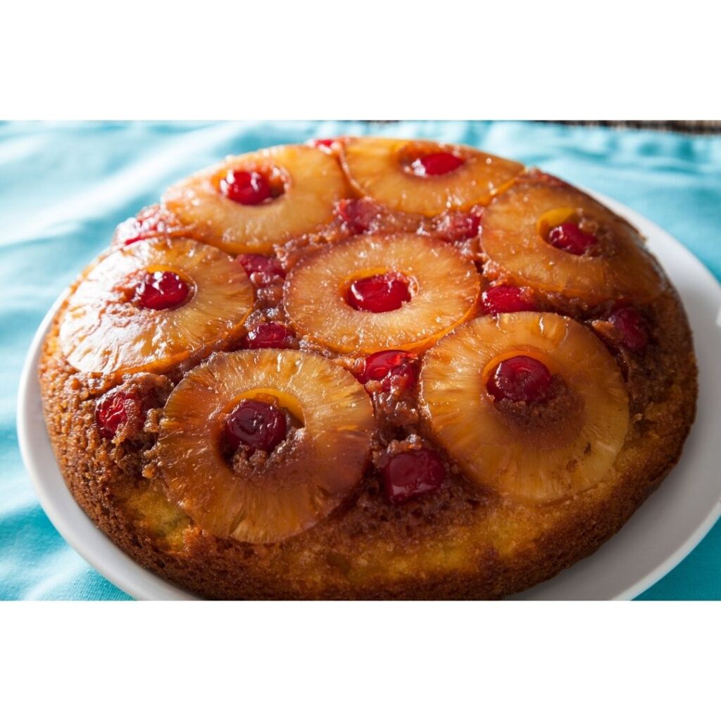 A cake with pineapple rings and maraschino cherries baked into the top, on a white plate on a blue table cloth.