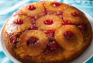 A cake with pineapple rings and maraschino cherries baked into the top, on a white plate on a blue table cloth.