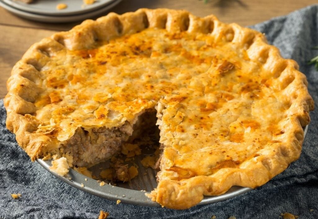 A tourtiere in a metal dish. The upper pastry is a golden brown and the edge is crimped. There is a piece missing.