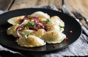 Several pierogis on a black plate with onions on top. There is a blue-gray piece of fabric on a wooden surface underneath the plate