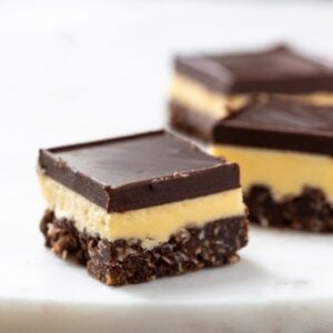 Three Nanaimo bars on a white plate. The bars have a crumbly chocolate base, then a yellow custard layer, with a final layer of chocolate on top. 