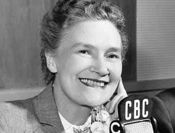 A black and white headshot of a woman in a suit jacket smiling in front of a microphone marked "CBC". Her hair is up and her hand is resting on the side of her face.