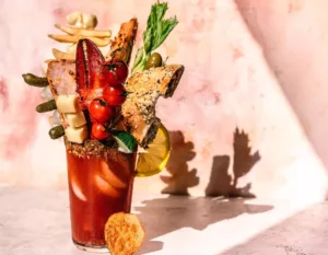 A Caesar in a glass, the beverage is red and has a dark brown rimmer. The glass is completely full of garnishes including a bagel, a lobster claw, cheese, french fries and tomatoes