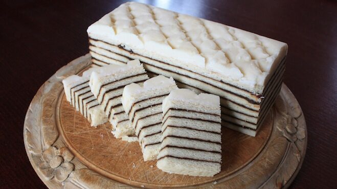 A rectangular cake with thin alternating layers of light coloured cookie and a dark purple filling with a white frosting on top. There are four slices of the cake infront of the unsliced portion, it is on a circular wooden serving platter.